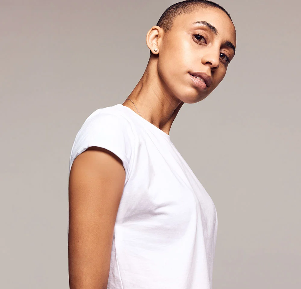 A person with a buzz cut and a white t-shirt looks over their shoulder directly at the camera against a plain gray background. - Abdominal Phalloplasty Abdophallo in Miami, Fl