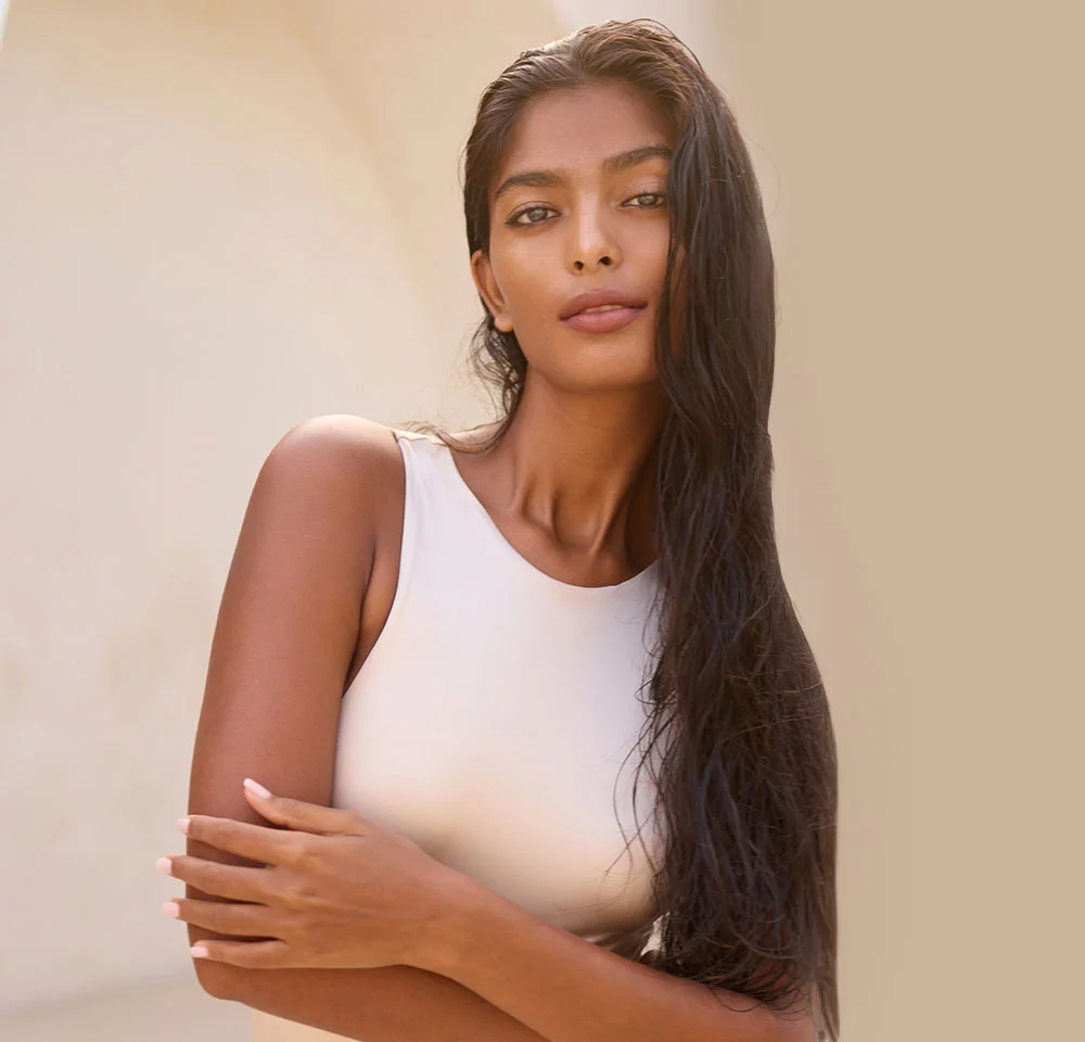 A woman with long, dark, wet hair is looking directly at the camera, wearing a high-necked, light beige or white top. She is standing against a warm, light beige background and has her arms crossed in front of her chest. - Arm Lift in Miami, Fl
