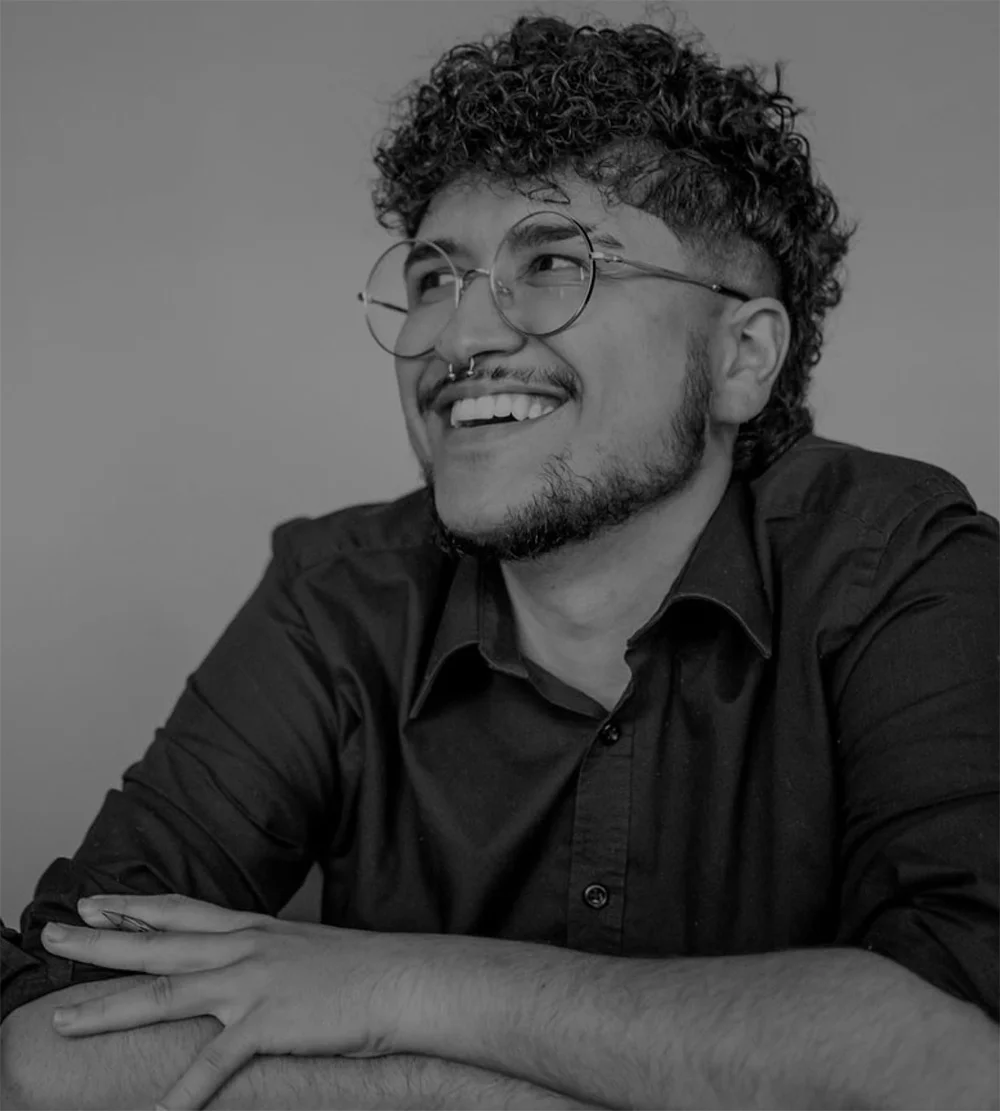 Black and white portrait of a young person with curly hair, a light beard, and glasses, smiling broadly and looking off to the side. They are wearing a dark collared shirt and have a septum piercing. Their arms are crossed on a surface in front of them.