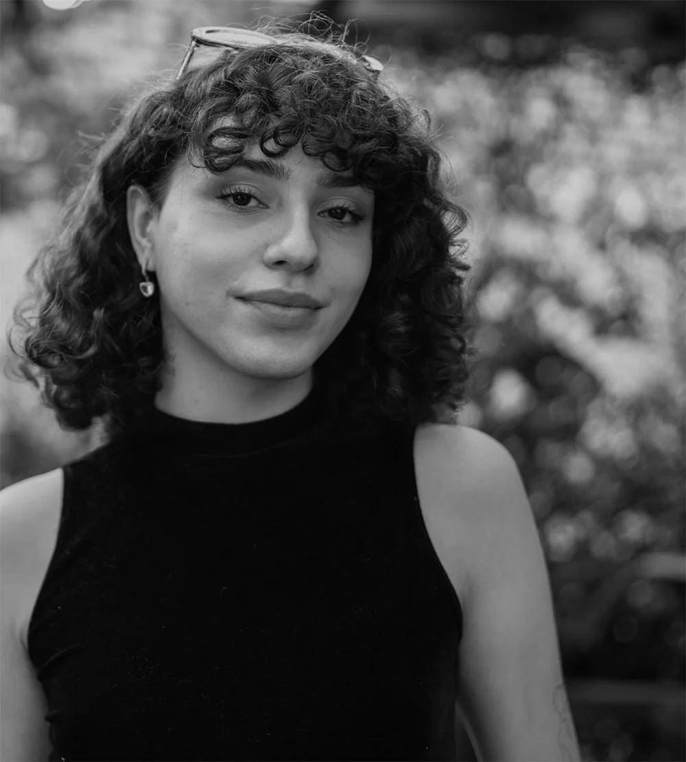 Black and white portrait of a young person with curly, medium-length hair and small hoop earrings, wearing a black, sleeveless mock-neck top. They are looking directly at the camera with a neutral expression. Sunglasses are perched on top of their head.