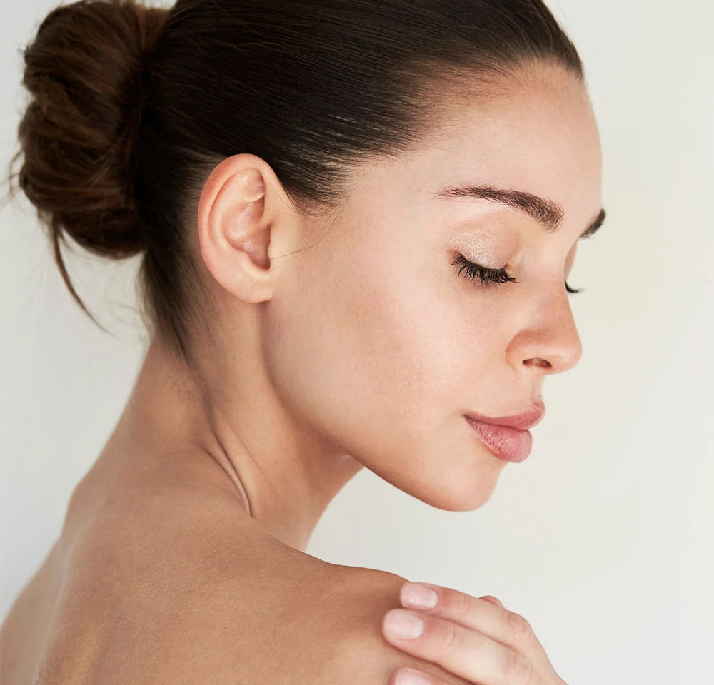 A close-up side profile of a woman with her eyes closed and her hair pulled back into a bun. She has natural makeup and is bare-shouldered, gently touching her right shoulder with her right hand. The background is a soft, light color. - Buffalo Hump Reduction in Miami, Fl