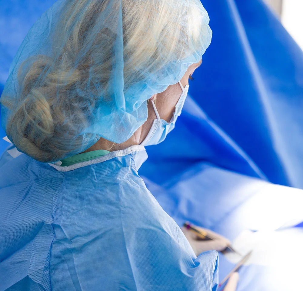 Dr. Gallagher, wearing a surgical cap and mask, is shown from the back of the head and shoulder, dressed in a blue surgical gown, looking down toward a bright operating area. - Double Incision Top Surgery in Miami, Fl