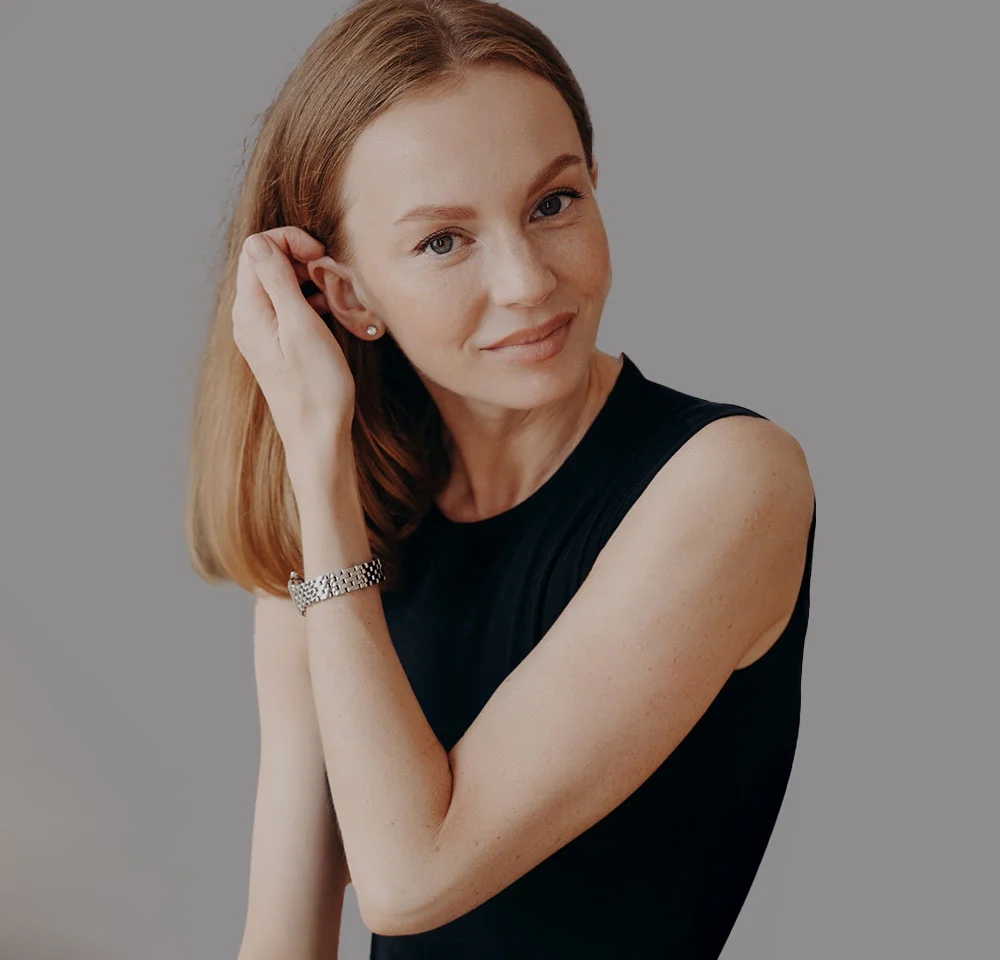 A woman with shoulder-length reddish-blonde hair and light freckles wears a black sleeveless top, a silver watch, and a stud earring, gently holding her hair back from her ear while looking at the camera against a grey background. - Elbow Lift in Miami, Fl