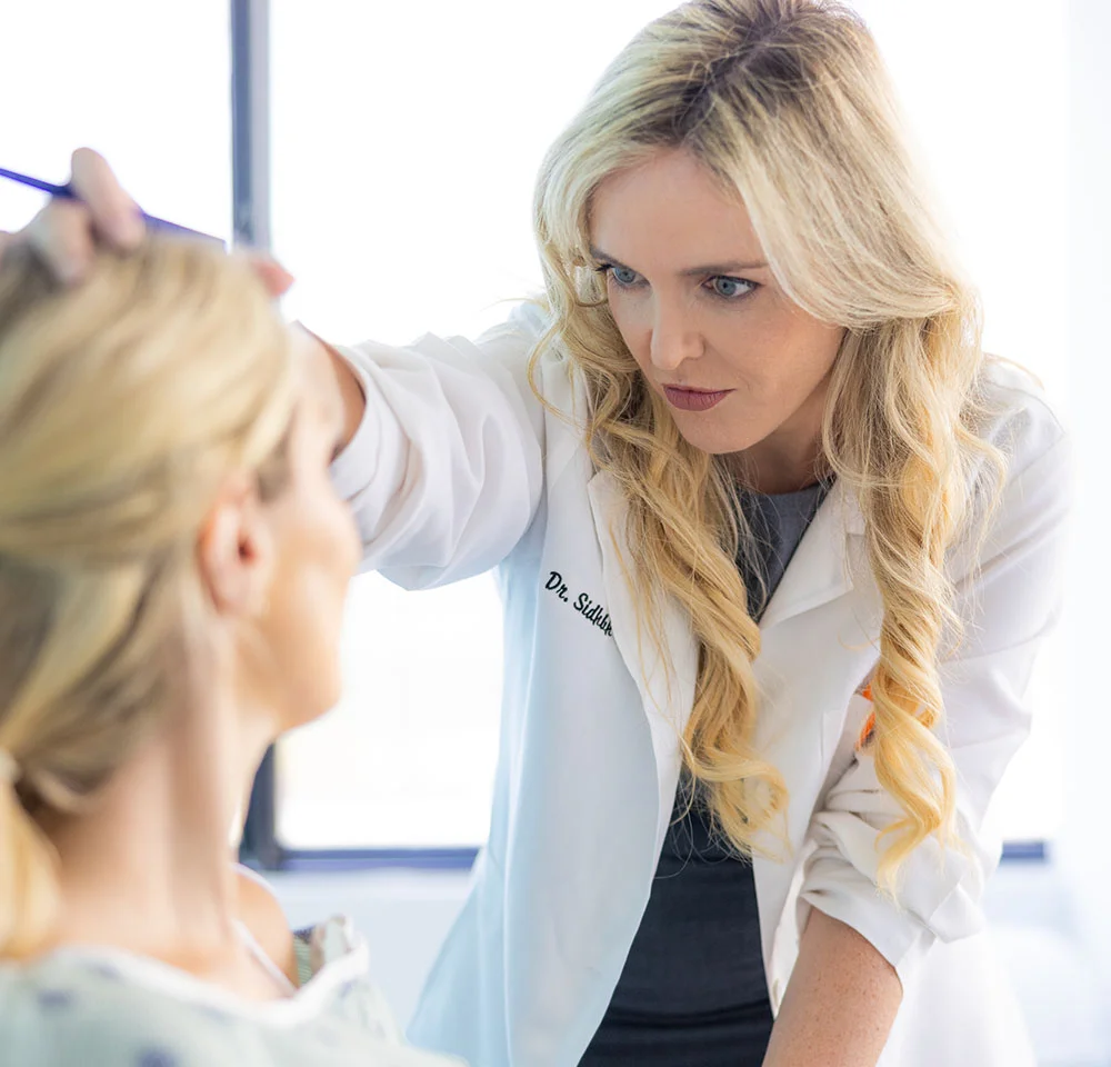 Dr. Gallagher holding a thin blue instrument, possibly a measuring tool, near the patient's forehead. The patient, whose back is to the camera, has blonde hair and is wearing a light-colored gown. The scene is bright, suggesting a medical examination room. - Forehead Reduction in Miami, Fl