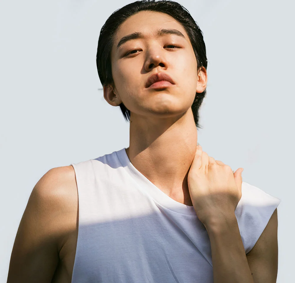A young East Asian man with dark, slicked-back hair wears a white sleeveless top. He looks directly at the camera with a serious expression while resting his left hand on the front of his neck. Sunlight casts a harsh shadow across his left shoulder and arm. - Gender Affirming Arm Lift in Miami, Fl