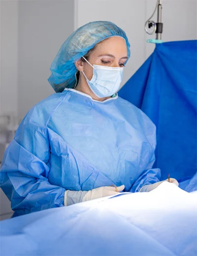 Dr. Ghallager, wearing a light blue surgical gown, a hair net, and a face mask, is focused on a procedure in an operating room. They are holding surgical instruments and looking down at the patient area covered by blue drapes.