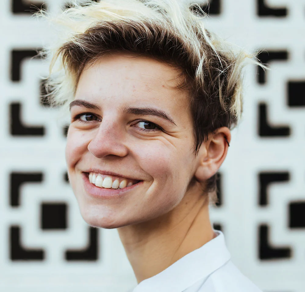 A close-up portrait of a young person with short, spiked hair dyed blonde on top, smiling broadly at the camera. They are wearing a white collared shirt. The background is a graphic black and white wall pattern made of repeating square designs. - Labiaplasty in Miami, Fl