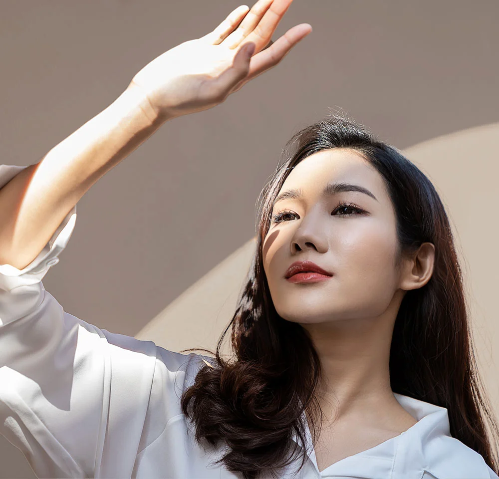 A close-up portrait of a young Asian woman with long dark hair, wearing a white flowing top. She is standing outdoors or near a bright window, shielding her eyes from the bright sunlight with her raised hand, and looking up and away. The background is a plain warm, beige color with a curved shadow pattern. - Lip Lift in Miami, Fl