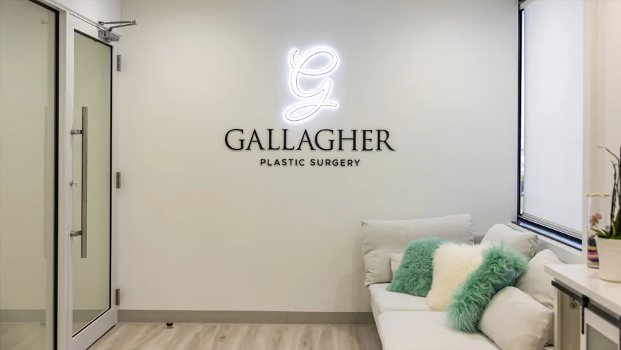 Interior view of the entrance and reception area of a plastic surgery office, showing a white wall with a neon 'G' and the "GALLAGHER PLASTIC SURGERY" logo in black lettering, next to a glass door and a white seating bench with pillows.