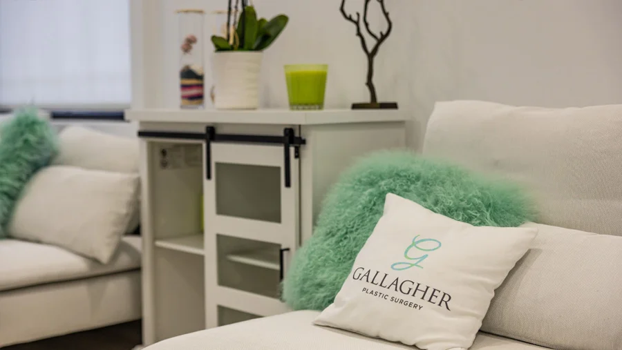 Interior view of a clean, bright waiting room featuring a white sofa with decorative pillows, including one with the "Gallagher Plastic Surgery" logo, and a white side cabinet with decorative plants and objects.