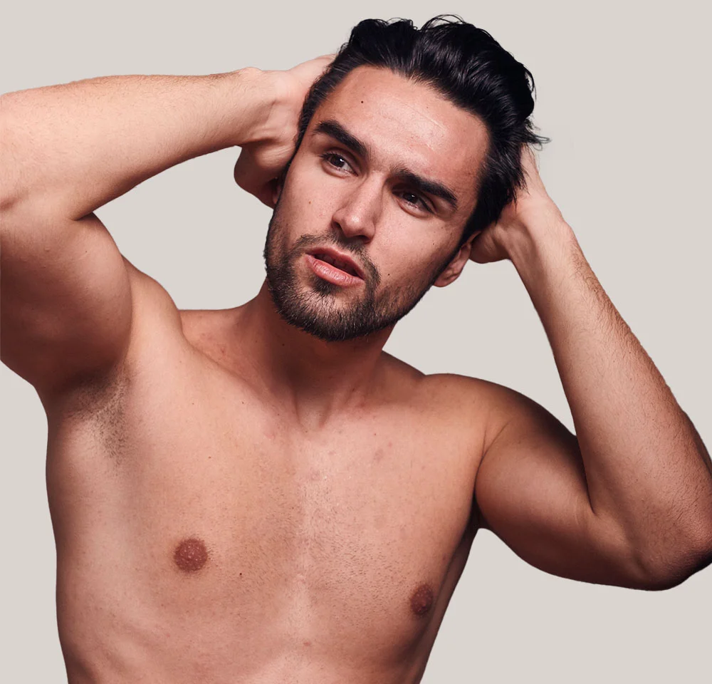 A handsome man with dark hair and a beard is shirtless and looking off to the side with an intense expression. He has both hands resting behind his head, partially running his fingers through his hair. The background is a solid, neutral gray or beige color. - Male Breast Reduction in Miami, Fl