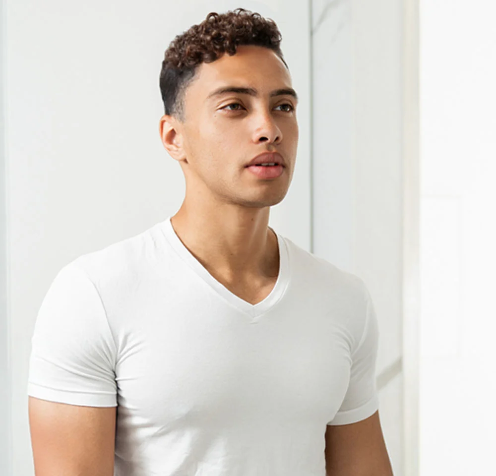 A young person with short, dark, curly hair and light eyes is shown from the chest up, looking off to the side. They are wearing a plain white V-neck t-shirt. The background is bright white, possibly a bathroom, with a faint vertical marbled detail on the wall behind them. They have a serious or contemplative expression. - Masculinizing Body Lift in Miami, Fl