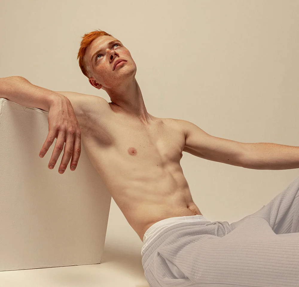 A pale, red-haired, and freckled, gender-non-conforming person is sitting down against a white background, leaning back on a white cube to the left. They are bare-chested and wearing light-colored, ribbed pants. They have a lean physique and are looking up and to the right with an open expression. - Masculinizing Tummy Tuck in Miami, Fl