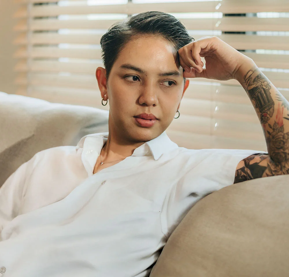 A person with short, slicked-back dark hair and a few visible arm tattoos is seated on a light-colored couch. They are wearing a white button-down shirt, silver hoop earrings, and are resting their hand near their head while looking thoughtfully off to the side. Light filters in from blinds in the background. - Metoidoplasty in Miami, Fl