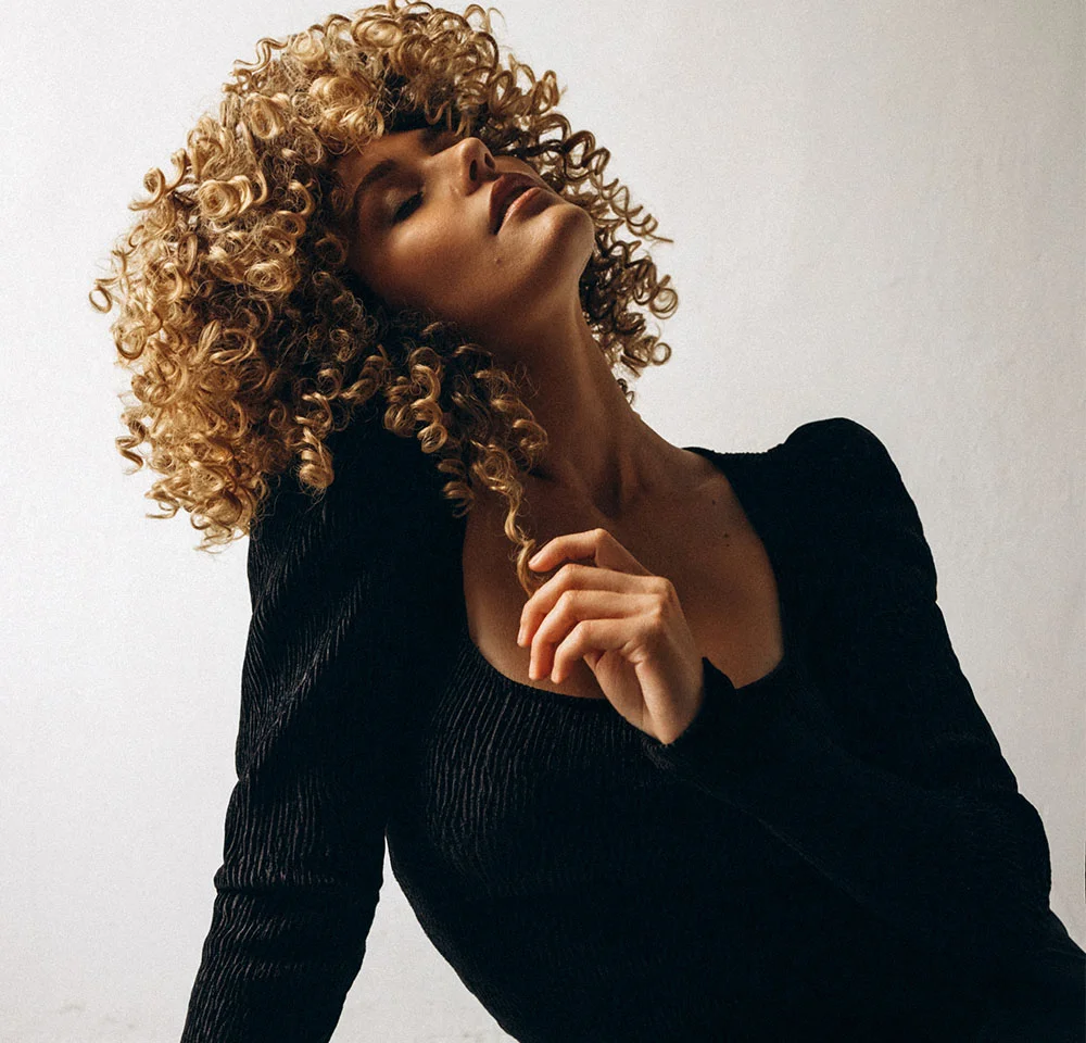 A close-up portrait of a woman with voluminous blonde curly hair, wearing a black textured top. She is looking at the camera while gently touching her chin and neck. - Neck Roll Removal in Miami, Fl