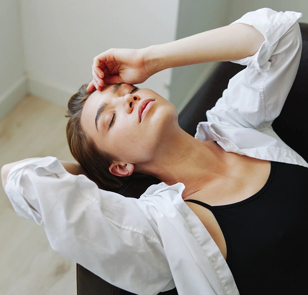 A close-up, high-angle shot of a person with short, dark hair lying on a dark surface, possibly a couch. Their eyes are closed, and they have a peaceful expression. They are wearing a black tank top with an open white button-down shirt draped over their shoulders and arms. One arm is resting above their head and the other is bent over their forehead. The background is a bright, white wall. - Non-Flat Top Surgery in Miami, Fl