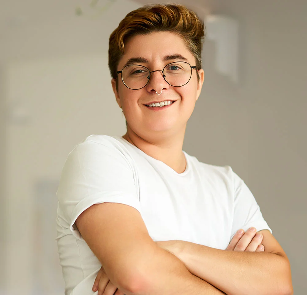 A person with short, reddish-brown hair and round glasses smiles while looking directly at the camera. They are wearing a simple white t-shirt and have their arms crossed. A small nose piercing is visible. The background is blurred and light gray. - Phalloplasty in Miami, Fl