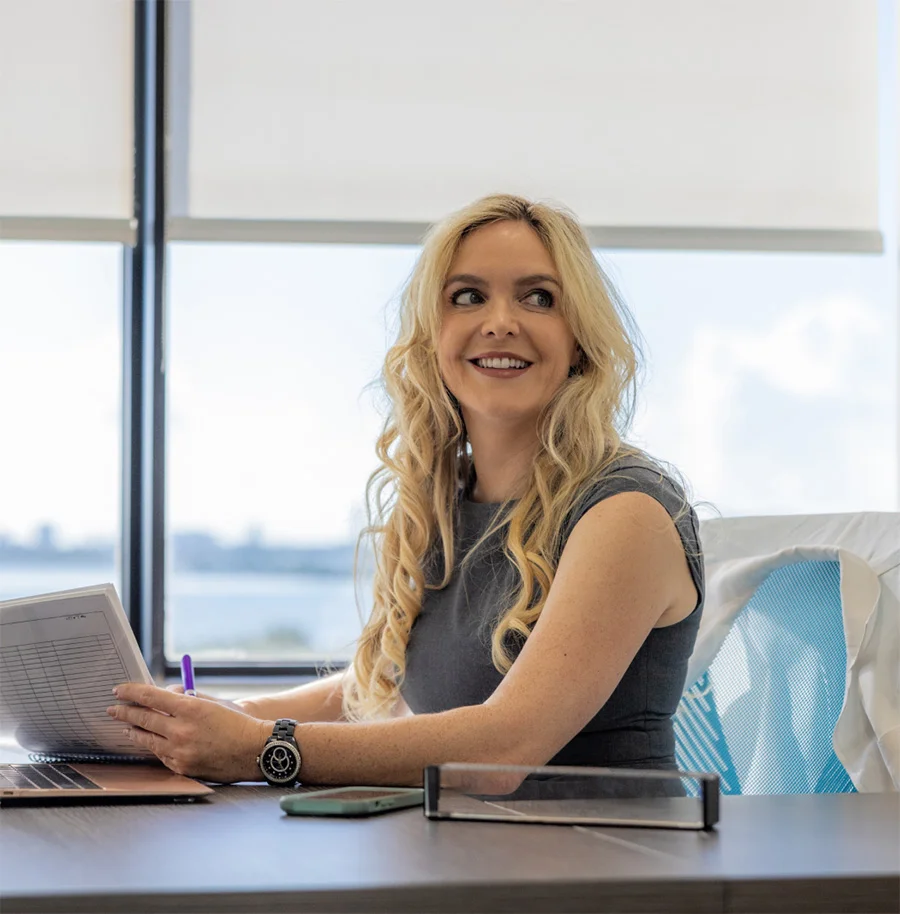 Dr. Gallagher, a blonde woman with long curly hair, is seated at a desk in her office with a large window overlooking a cityscape. She is smiling and looking off to the side while holding documents and a pen.