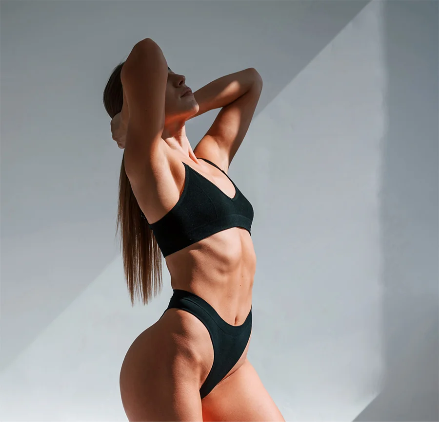 A toned woman with long brown hair stands in profile, wearing a black sports bra and briefs. One hand rests near her head, her expression serene. Bright lighting casts soft shadows on a plain light gray wall, highlighting her smooth, tanned physique.