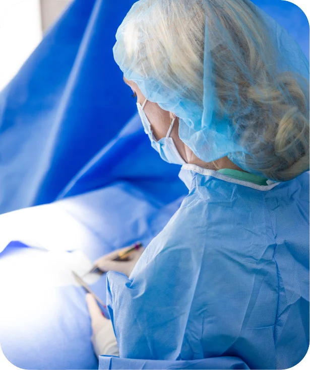 Dr. Gallagher, wearing a surgical cap and mask, is shown from the back of the head and shoulder, dressed in a blue surgical gown, looking down toward a bright operating area.