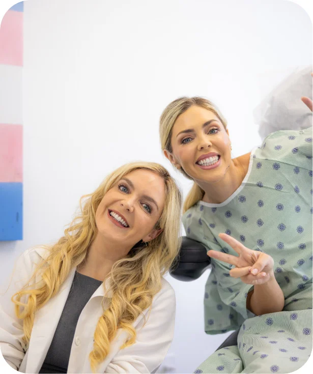 Dr. Gallagher (left), in a white lab coat, and a blonde patient (right), in a blue patterned hospital gown, smile and look up at the camera. The patient is holding up a peace sign with one hand. A piece of art with the colors of the transgender flag is visible on the wall behind them.