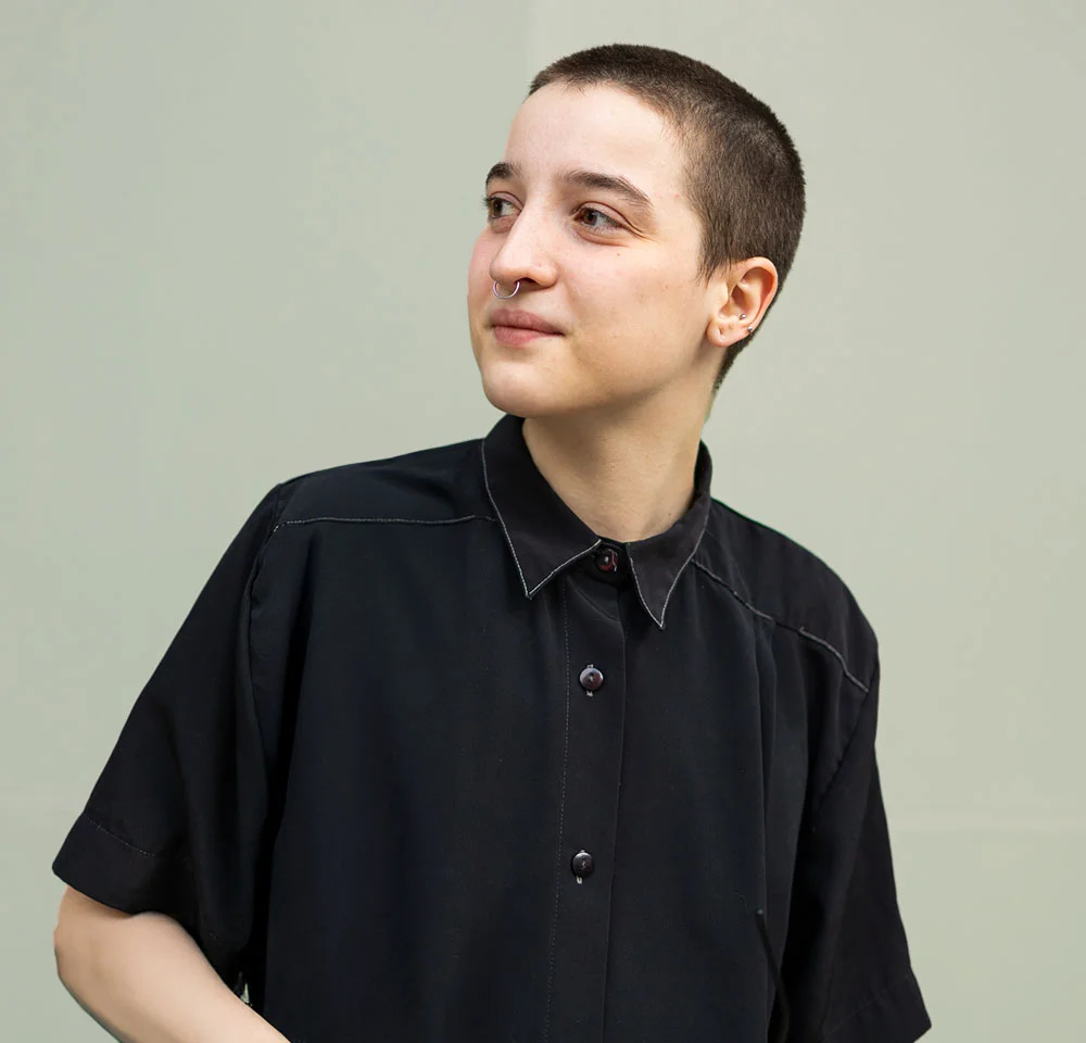 A person with a short buzz cut and a small nose ring looks toward the upper right, away from the camera. They are wearing a short-sleeved black collared shirt with visible white stitching. The background is a plain light greenish-gray. - Scrotoplasty in Miami, Fl