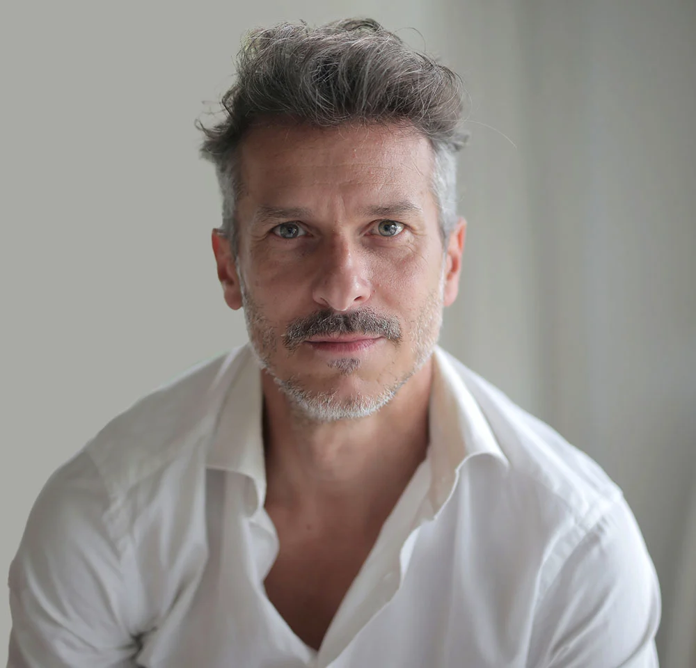 A middle-aged man with graying hair and a mustache looks directly at the camera. He is wearing a white collared shirt that is open at the neck, set against a plain light gray or white background. - Scrotum Lift in Miami, Fl