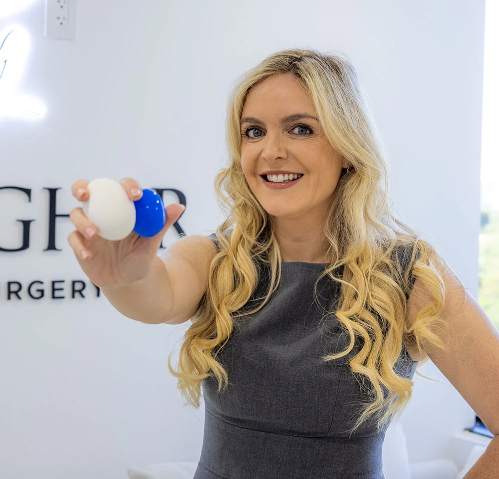 Dr. Gallagher, smiling with long blonde wavy hair, wearing a gray sleeveless dress, holds two egg-shaped objects (one white, one blue) toward the camera. Behind her, part of a wall with the stylized words "GALLAGHER SURGERY" is visible in the background. - Testicular Enhancement in Miami, Fl