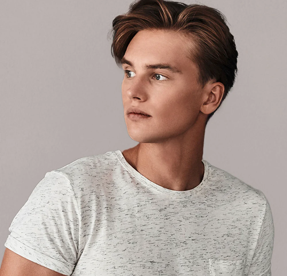 A close-up headshot of a man with short brown hair, a slight beard, and light blue eyes, wearing a white long-sleeved shirt, looking off to the left against a neutral gray background. - Top Surgery in Miami, Fl