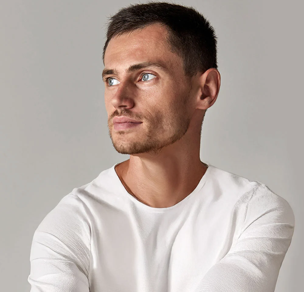 A close-up headshot of a man with short brown hair, wearing a white long-sleeved shirt, looking off to the left against a neutral gray background. He has light blue eyes and a slight beard. - Top Surgery with Tummy Tuck in Miami, Fl
