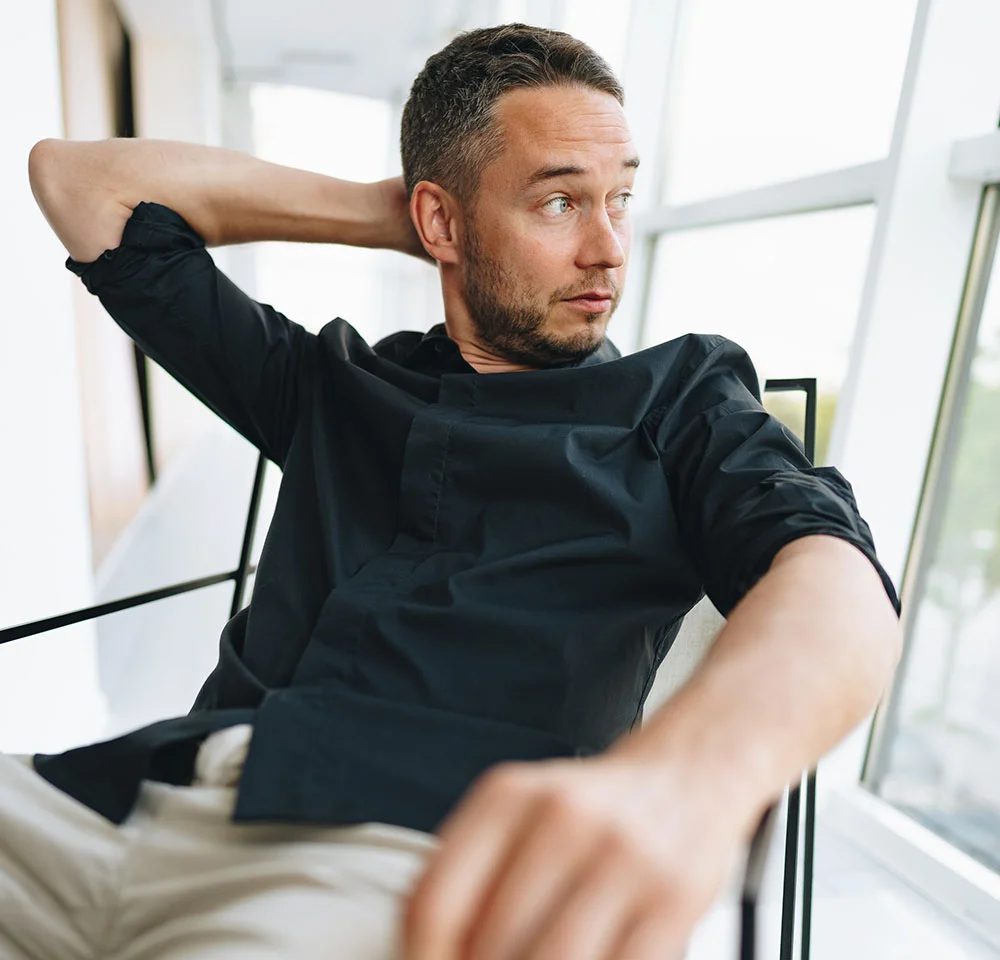 A man with short hair and a beard, wearing a black button-down shirt and light-colored pants, is seated, looking off to the side. He has his arms crossed behind his head. The background is a bright, modern interior with large windows. - Torso Masculinization in Miami, Fl