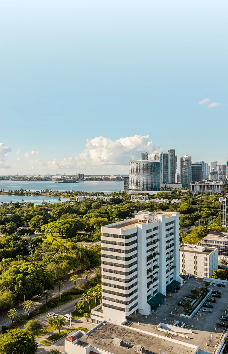 Vertical aerial view of a Miami cityscape with lush green trees, a prominent white office building in the foreground, and high-rise buildings along the bay under a bright blue sky.